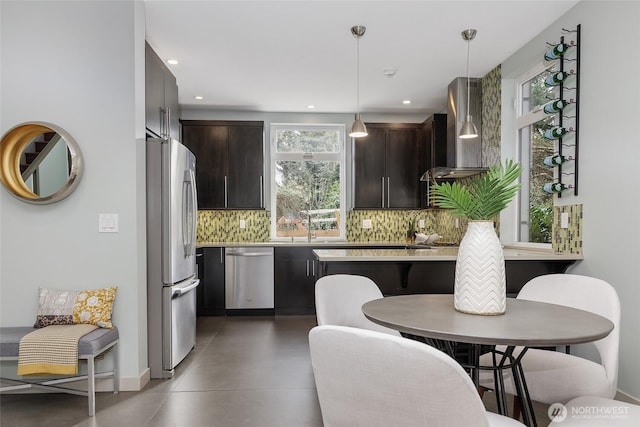 kitchen with stainless steel appliances, dark brown cabinets, wall chimney exhaust hood, and decorative light fixtures