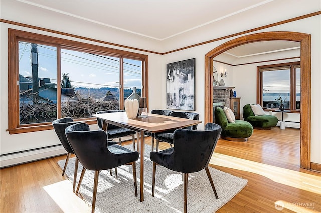 dining space with arched walkways, light wood-style flooring, a fireplace, baseboard heating, and crown molding