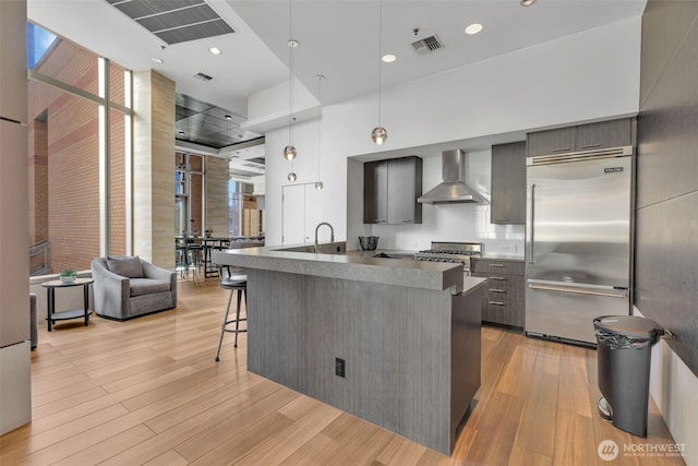 kitchen with pendant lighting, light hardwood / wood-style flooring, built in refrigerator, a kitchen bar, and wall chimney exhaust hood