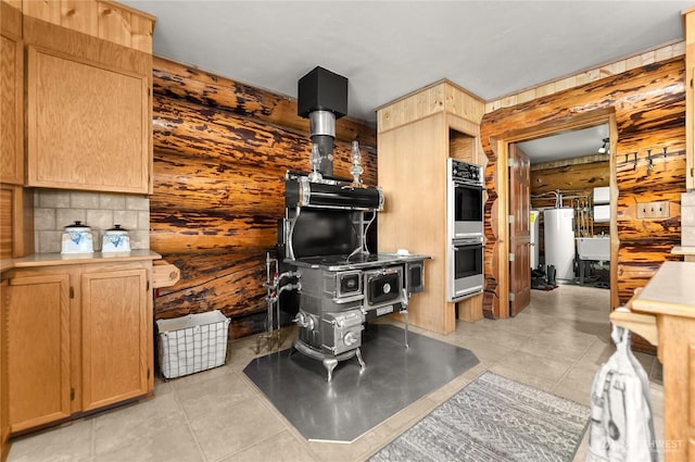 kitchen featuring double wall oven, light brown cabinetry, gas water heater, wood walls, and a wood stove