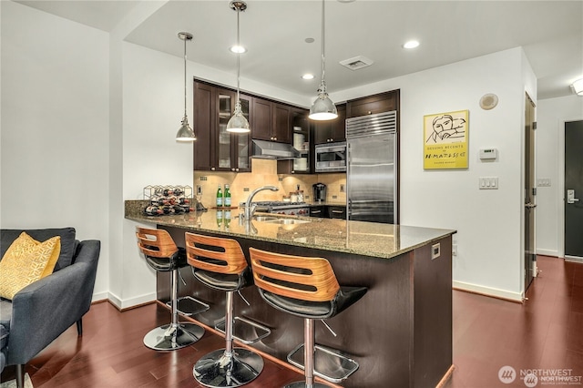 kitchen with dark brown cabinetry, a kitchen bar, built in appliances, dark stone countertops, and kitchen peninsula