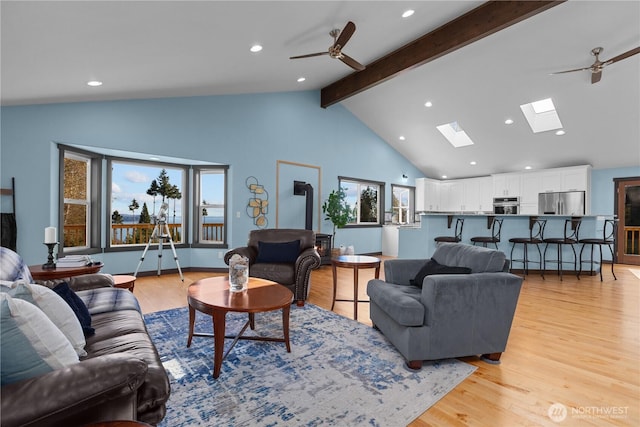 living room with a skylight, beamed ceiling, a wood stove, ceiling fan, and light hardwood / wood-style flooring