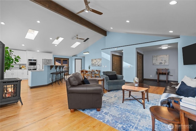 living room featuring ceiling fan, vaulted ceiling with skylight, light hardwood / wood-style floors, and a wood stove