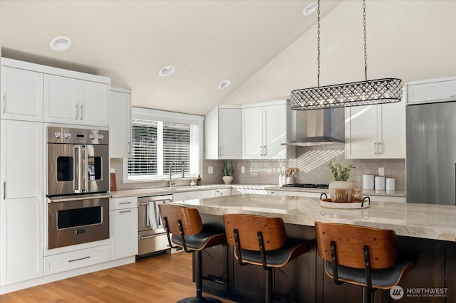 kitchen featuring wall chimney exhaust hood, light wood-style flooring, a breakfast bar, vaulted ceiling, and stainless steel appliances