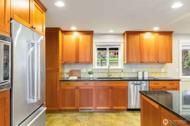 kitchen with brown cabinets, stainless steel appliances, backsplash, a sink, and dark stone counters