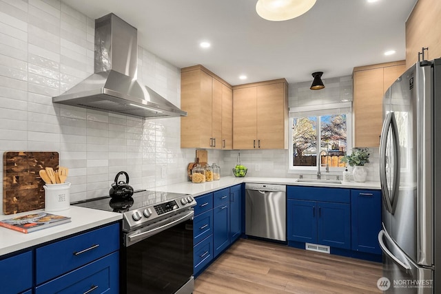 kitchen featuring wall chimney range hood, sink, appliances with stainless steel finishes, wood-type flooring, and blue cabinets