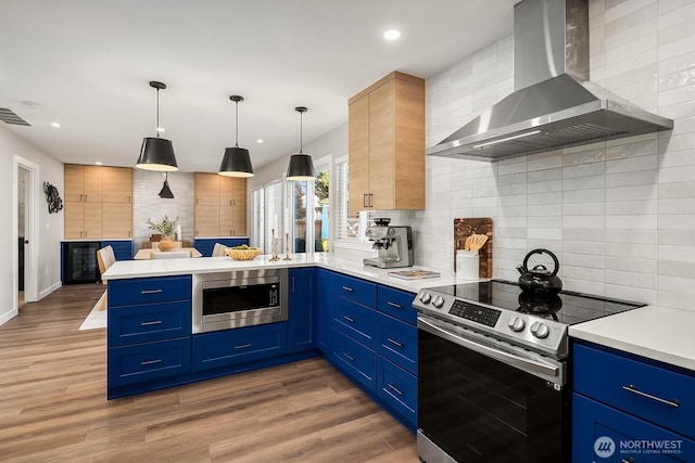 kitchen featuring wall chimney range hood, blue cabinetry, appliances with stainless steel finishes, decorative light fixtures, and kitchen peninsula