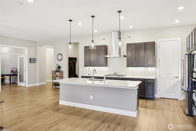 kitchen with dark brown cabinetry, wall chimney range hood, a kitchen island with sink, and stainless steel gas stovetop