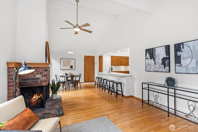 living room featuring light wood finished floors, a brick fireplace, vaulted ceiling with beams, baseboards, and a ceiling fan