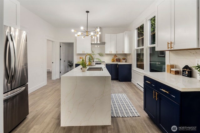 kitchen with blue cabinetry, hanging light fixtures, stainless steel fridge, a kitchen island with sink, and white cabinets