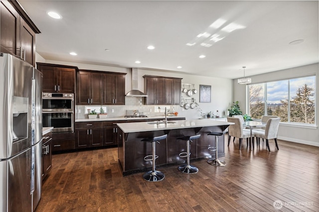 kitchen featuring hanging light fixtures, appliances with stainless steel finishes, light countertops, wall chimney exhaust hood, and an island with sink