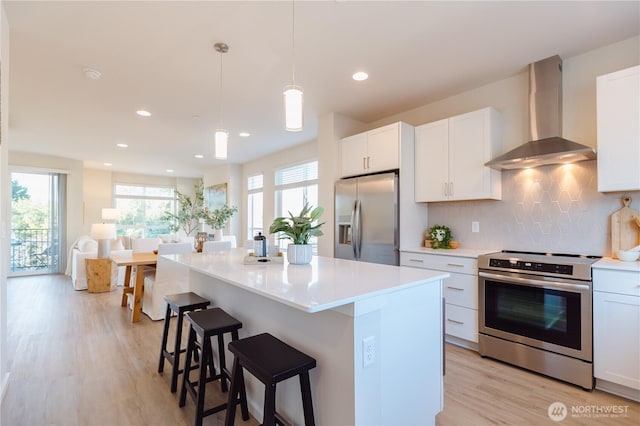 kitchen featuring wall chimney range hood, hanging light fixtures, stainless steel appliances, a center island, and white cabinets