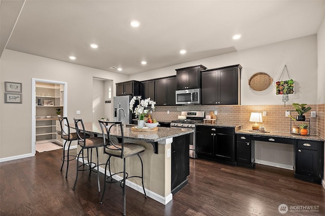 kitchen featuring dark wood-type flooring, a kitchen bar, light stone counters, an island with sink, and stainless steel appliances