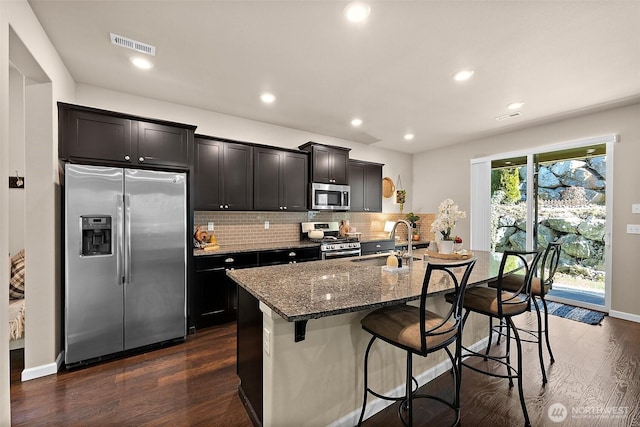 kitchen with a breakfast bar, sink, a center island with sink, dark stone countertops, and appliances with stainless steel finishes