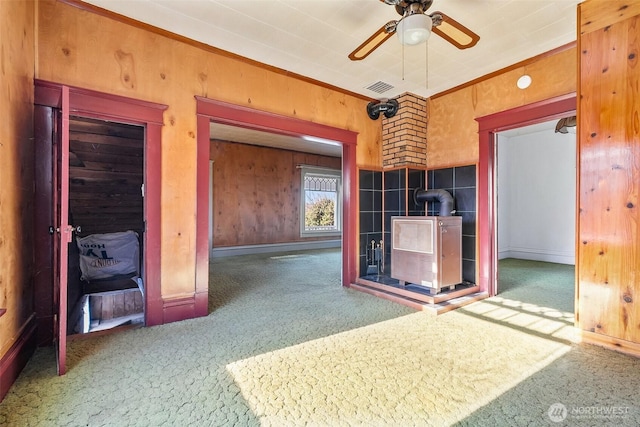 unfurnished living room with wood walls, a wood stove, carpet, ceiling fan, and crown molding