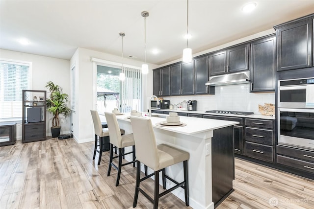 kitchen featuring pendant lighting, a center island with sink, stainless steel appliances, light countertops, and under cabinet range hood