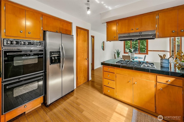 kitchen with under cabinet range hood, light wood-style floors, appliances with stainless steel finishes, brown cabinets, and dark countertops