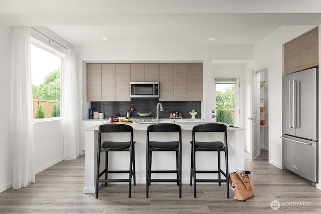 kitchen with a breakfast bar area, light wood-type flooring, stainless steel appliances, a kitchen island with sink, and backsplash