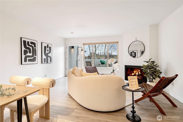 living room with a warm lit fireplace, light wood-type flooring, and baseboards