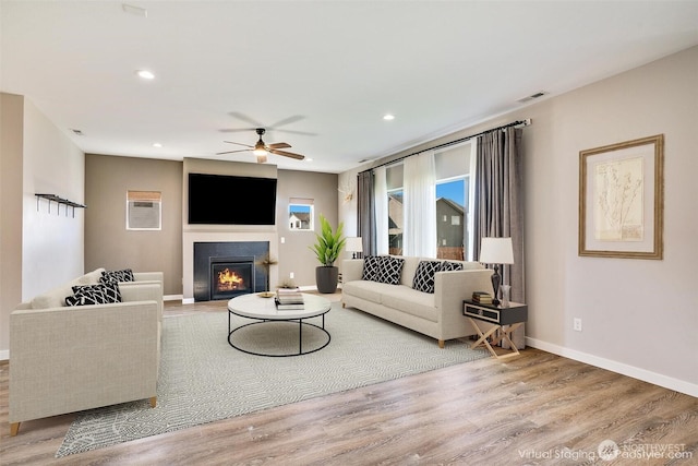 living area featuring a glass covered fireplace, a ceiling fan, baseboards, and wood finished floors