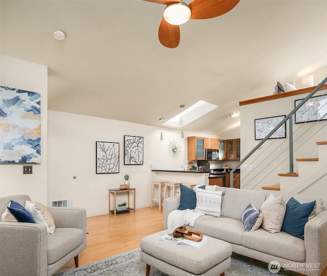 living area featuring light wood finished floors, visible vents, a ceiling fan, lofted ceiling with skylight, and stairway