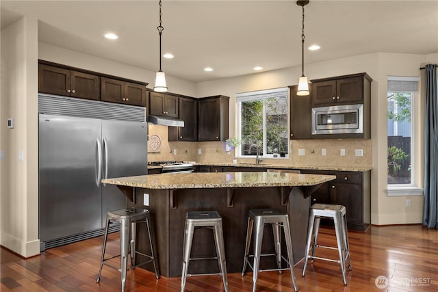 kitchen with dark wood-type flooring, under cabinet range hood, backsplash, dark brown cabinetry, and built in appliances