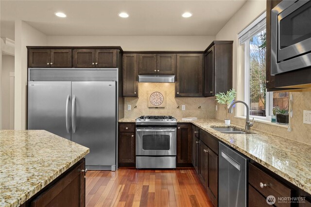 kitchen with hardwood / wood-style floors, a sink, dark brown cabinetry, under cabinet range hood, and built in appliances