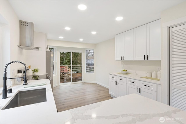 kitchen with freestanding refrigerator, white cabinets, a sink, wall chimney range hood, and light stone countertops