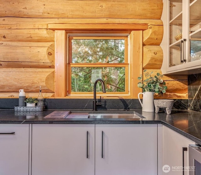 kitchen with log walls, tasteful backsplash, glass insert cabinets, white cabinetry, and a sink