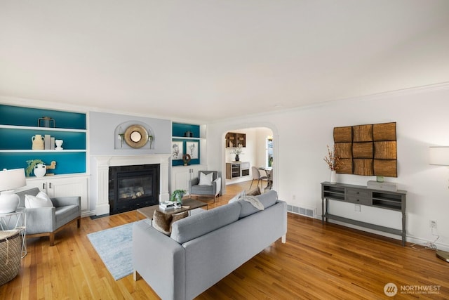 living area featuring built in features, visible vents, arched walkways, a glass covered fireplace, and wood finished floors