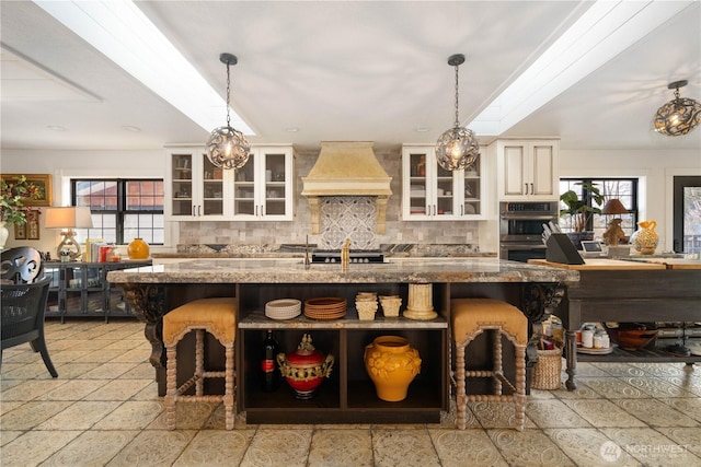 kitchen featuring premium range hood, stainless steel double oven, glass insert cabinets, and decorative light fixtures