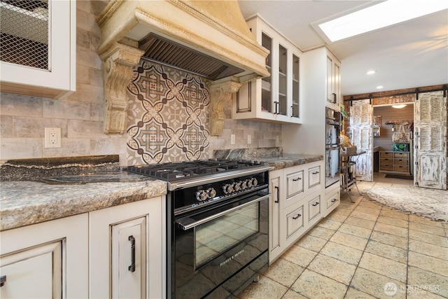 kitchen featuring decorative backsplash, glass insert cabinets, gas stove, dark stone countertops, and premium range hood