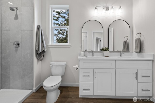 bathroom featuring toilet, double vanity, a tile shower, wood finished floors, and a sink