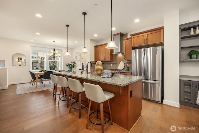 kitchen featuring an island with sink, light stone counters, wall chimney range hood, stainless steel refrigerator, and hanging light fixtures