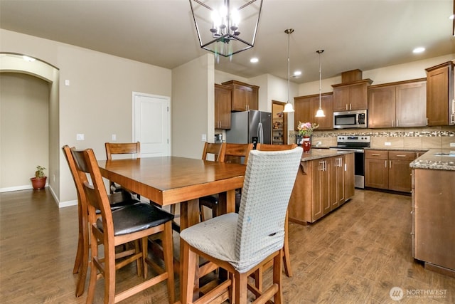 kitchen with light stone counters, hanging light fixtures, appliances with stainless steel finishes, brown cabinets, and dark wood-style floors