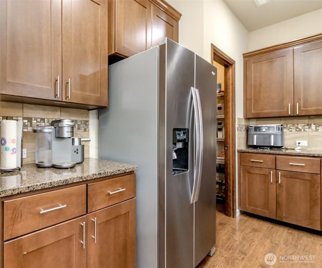 kitchen with stainless steel refrigerator with ice dispenser, light wood-style flooring, decorative backsplash, brown cabinetry, and light stone countertops