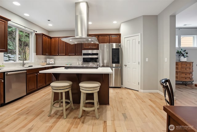 kitchen featuring island range hood, stainless steel appliances, light countertops, backsplash, and light wood finished floors