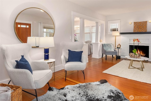 living area with a wealth of natural light, a brick fireplace, decorative columns, and wood finished floors