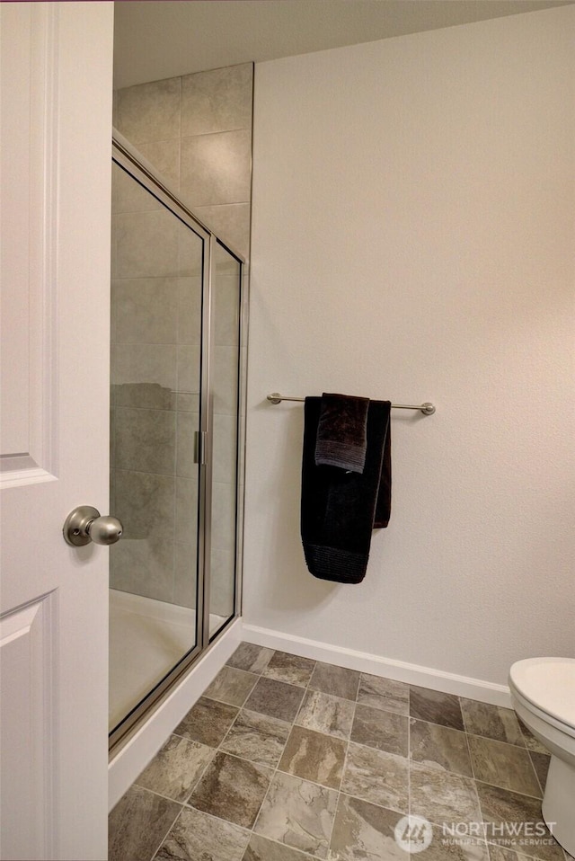 bathroom featuring stone finish floor, toilet, baseboards, and a stall shower