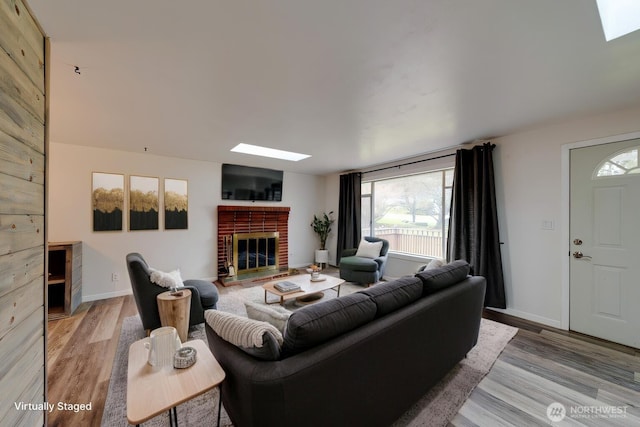 living area featuring a skylight, light wood-style flooring, a brick fireplace, and baseboards