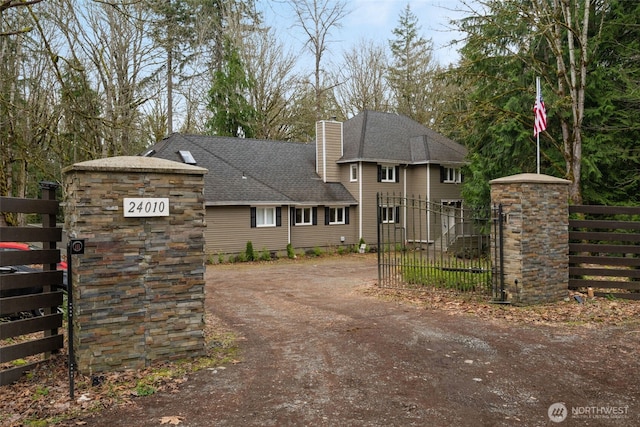 view of front facade featuring a gate, dirt driveway, fence, roof with shingles, and a chimney