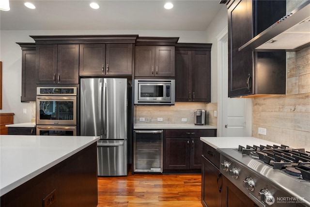 kitchen featuring wine cooler, stainless steel appliances, light countertops, dark brown cabinetry, and wall chimney range hood