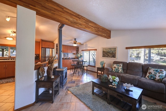 living room featuring vaulted ceiling with beams, ceiling fan, a textured ceiling, baseboards, and a wood stove