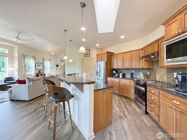 kitchen featuring backsplash, under cabinet range hood, open floor plan, a breakfast bar area, and appliances with stainless steel finishes