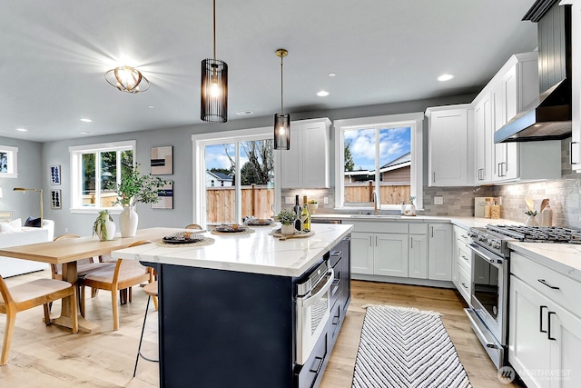 kitchen featuring stainless steel stove, a kitchen island, white cabinets, hanging light fixtures, and wall chimney range hood