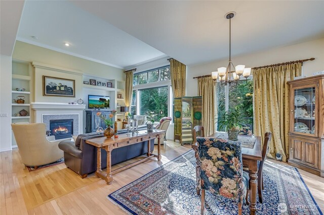 dining area with built in shelves, crown molding, light wood finished floors, an inviting chandelier, and a glass covered fireplace