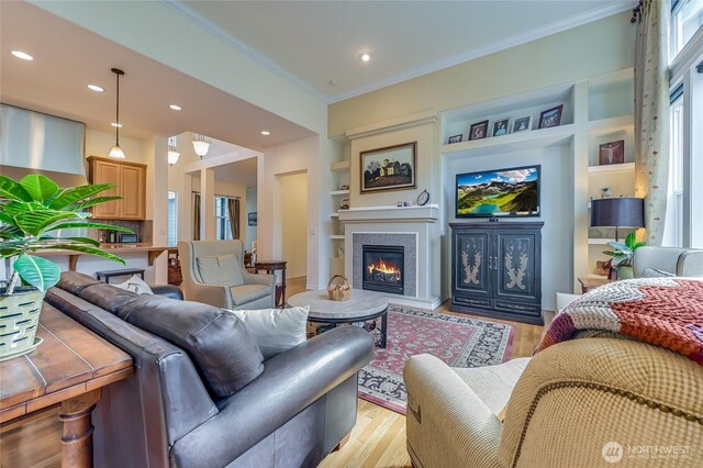 living area featuring built in shelves, recessed lighting, ornamental molding, light wood finished floors, and a glass covered fireplace