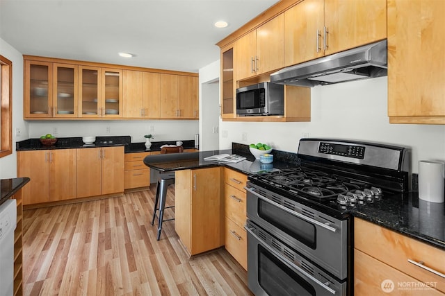 kitchen featuring appliances with stainless steel finishes, glass insert cabinets, dark stone countertops, light wood-type flooring, and under cabinet range hood