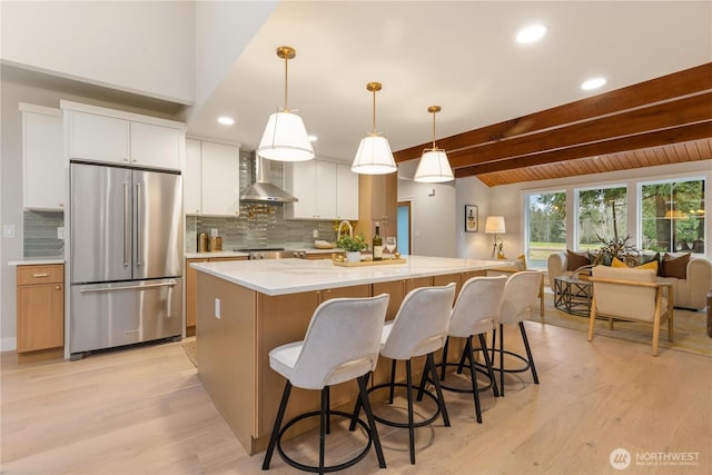 kitchen with white cabinets, an island with sink, high quality fridge, decorative light fixtures, and wall chimney range hood