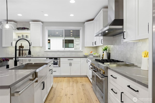 kitchen featuring light hardwood / wood-style flooring, wall chimney range hood, stainless steel appliances, white cabinets, and hanging light fixtures